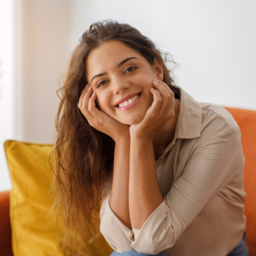 A woman with long brown hair, wearing a beige shirt, sits on an orange sofa and smiles at the camera with her hands resting on her face.