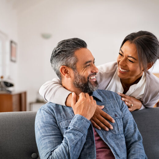 A man sits on a couch smiling at a woman who stands behind him with her arm on his shoulder; both appear happy in a bright living room.